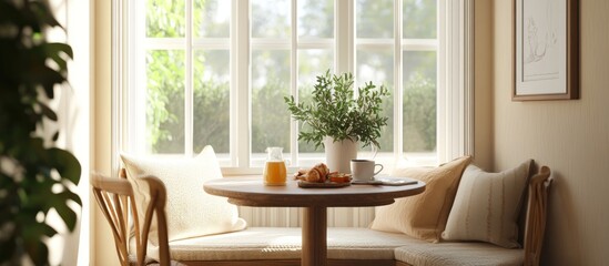 A cozy nook with a round table and two chairs by a window.  The table is set with a glass of orange juice, a croissant, and a mug.  There are pillows on the bench and a plant in a white pot.
