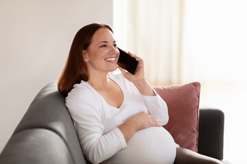 Obraz premium A smiling pregnant woman relaxes on a couch, cradling her belly while chatting on the phone. Natural light fills the cozy room, creating a warm atmosphere.