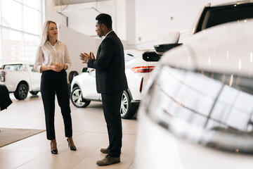 Full length portrait of female customer choosing auto, listening to African male dealer in auto dealership. Professional black car salesman telling thinking buyer about luxurious car in motor show.