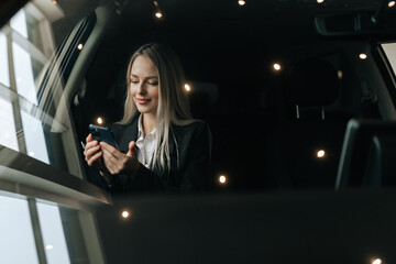 View through windshield to attractive blonde female in suit reading good news using mobile phone sitting on front passenger seat in car. Pretty young woman browsing smartphone, surfing internet.