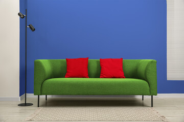 Stylish green sofa with red pillows and lamp near blue wall indoors