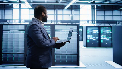 African american man walking in high tech facility, using laptop to patch network issues leading to slow data transfer rates. Data center worker fixing errors affecting supercomputers, camera B