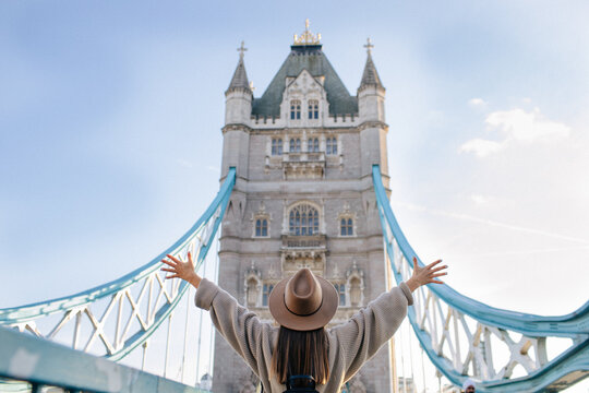 Young beautiful tourist girl is happy and excited to see Tower bridge in London, she is wearing a hat and raised her hands up, the view from a back 