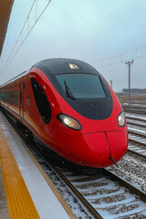 Fototapeta premium A sleek red train waits at a train station, surrounded by a snowy landscape on a chilly winter day with overcast skies