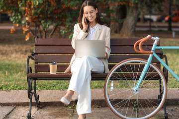 Young businesswoman with bicycle and laptop talking by phone while sitting on bench in park