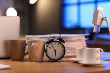Alarm clock with papers and cups of coffee on table in office at night, closeup. Deadline concept