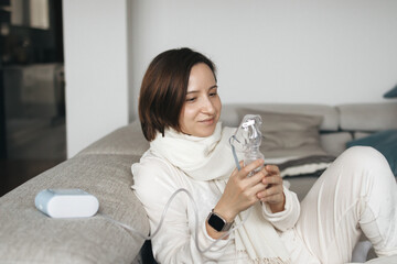 Young Woman Is Sitting On A Sofa At Home, Using A Nebulizer To Help Her Breathe