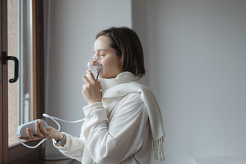 Young Woman Is Standing By The Window, Breathing From A Portable Oxygen Concentrator