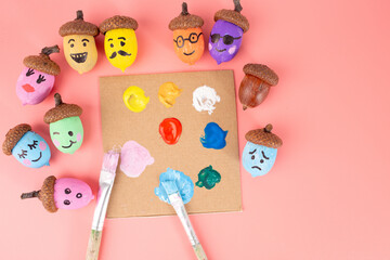 painted acorns are arranged on pink background. Additionally, there is a small palette with blobs of paint in various colors and two paintbrushes with paint on them.