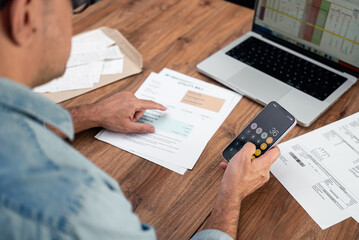 Mature businessman adds utility bills numbers with smartphone app at office desk, senior male...