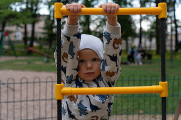 Fototapeta premium Boy on the playground hanging on the stairs wearing a dinosaur-themed sweatshirt in the park on a calm day. Concept of children's play, outdoor fun and healthy physical activity. High quality photo