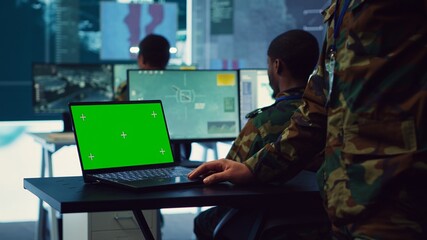 Army officer studies isolated greenscreen display on a laptop in control room, military power base...