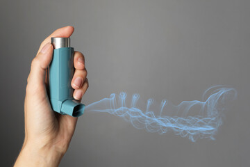 Asthma treatment. Woman holding inhaler on grey background, closeup. Aerosolized medication coming...