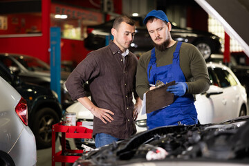 Positive young auto-mechanic in blue overalls communicating with male client of car workshop about...