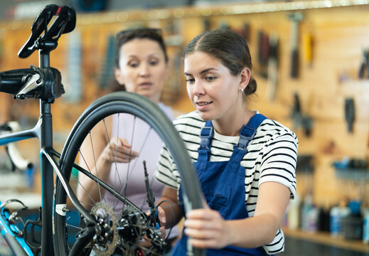 Professional confident young female bicycle technician adjusting bike gear system, fixing sprockets on rear wheel using cassette pliers and explaining repair details to female client in workshop