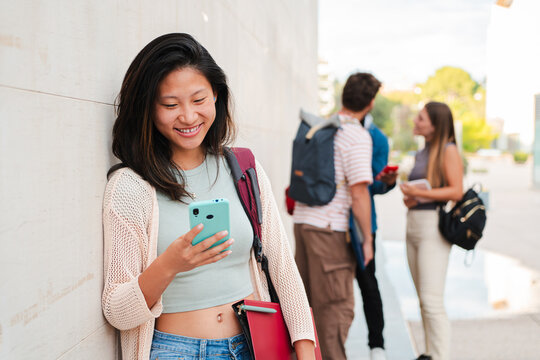 Young chinese girl smiling while looking at her cellphone, leaning against a wall outside. Casual moment of a student or teenager using mobile technology to text, browse, or connect with friends