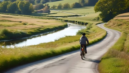 Obraz premium A serene journey through nature a bicyclist pedaling along a winding riverbank road surrounded by picturesque green fields and trees under a clear blue sky