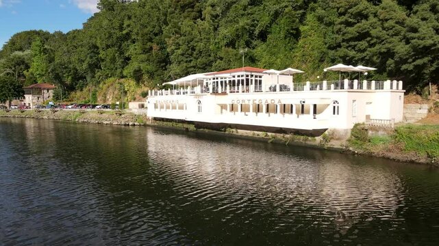 A serene riverside scene featuring a modern health resort nestled along the water&rsquo;s edge. The building, painted in light tones, features an outdoor terrace with umbrellas and seating areas
