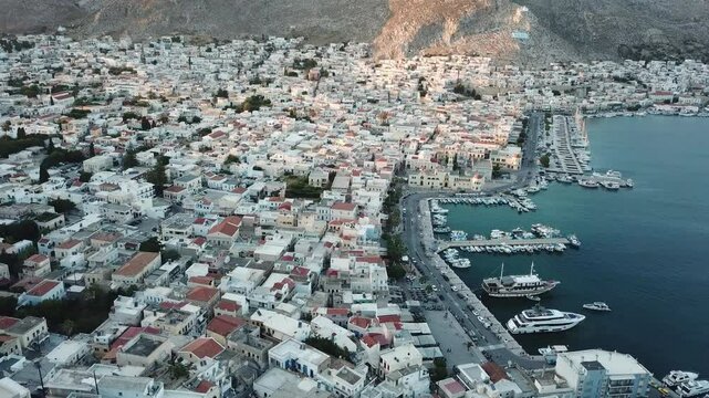 Greece,Kalimnos,Drone Footage high and above the harbor and the main town during sunset, sun is shinning on the steep hill of the town where the town is in shadow, lot of boats docked at the harbor.