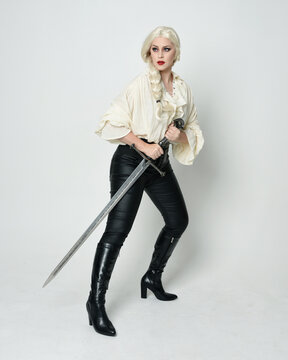 Full length portrait of beautiful female model wearing vintage gothic fantasy blouse, leather pants, boots and blonde plaited hair, larp costume.  standing pose housing sword weapon in fighting action