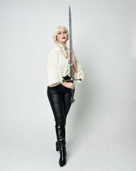 Fototapeta premium Full length portrait of beautiful female model wearing vintage gothic fantasy blouse, leather pants, boots and blonde plaited hair, larp costume. standing pose housing sword weapon in fighting action