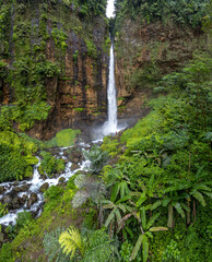 A someone stands at the base of the majestic Kapas Biru Waterfall in Indonesia. The stunning natural beauty of the waterfall is emphasized by the surrounding rainforest.