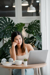 Fototapeta premium A young woman focused on her laptop in a stylish home office setting, surrounded by lush green plants and soft natural lighting.