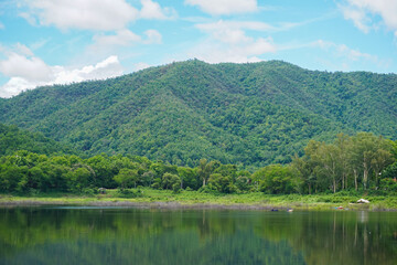 A beautiful nature landscape of the greenery mountains, blue sky, and clouds reflect on a calm water. A symbol of a relaxing holidays, nature, countryside, forest, camping site, calm, and stillness