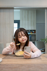 Asian girl enjoys watching cartoons on her smartphone while having breakfast and eating cereal at the dining table.