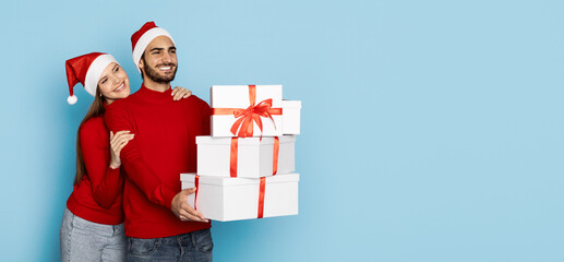 A cheerful couple wearing red sweaters and Santa hats poses together while holding beautifully wrapped gift boxes with red ribbons. The bright blue background, copy space