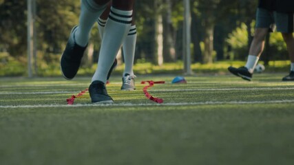 Low section of two unrecognizable boys wearing sports shoes and long white socks practicing agility drill on sunlit grassy soccer field during outdoor training with coach at daytime