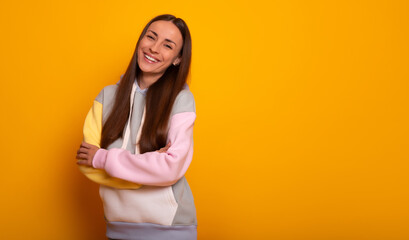 Obraz premium A cheerful young woman smiles confidently against a bright yellow background. Wearing a colorful hoodie with crossed arms, she exudes happiness and casual style in this vibrant half-body shot.