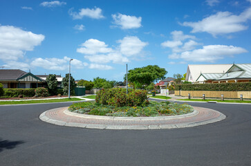 A roundabout is surrounded by residential homes with well-manicured lawns and fences on an Australian suburban street.