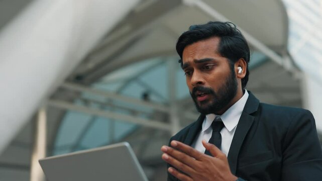 Nervous businessman in shirt gesturing while speaking during remote meeting sitting outside office building. Male manager looking at screen of computer while making excuses for mistake made