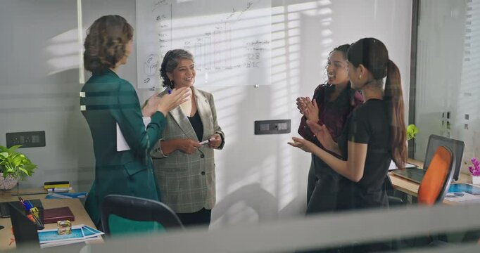 Indian happy old boss lady leader show growth chart graph data on white board share idea plan work place older female talk speak good news expert intern team girl group clap hands indoor job office