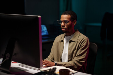 African American man concentrating on work in front of computer screen in dim office environment,...