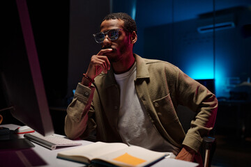Employee contemplating work in modern office, surrounded by open book, keyboard, and eyeglasses on desk. Soft blue lighting illuminating background with focused demeanor