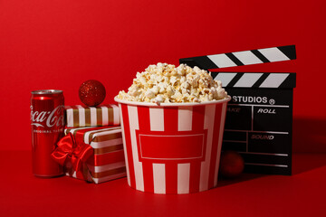 Large paper cup of popcorn, clapperboard and gift boxes on red background