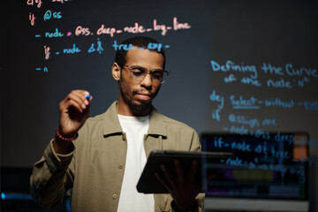 African American man concentrating on tablet while reviewing programming code on digital screen during software development session in office setting