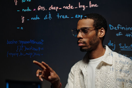 African American software developer working on a transparent screen displaying code. Concentrated look with focused expression and high-tech coding happening