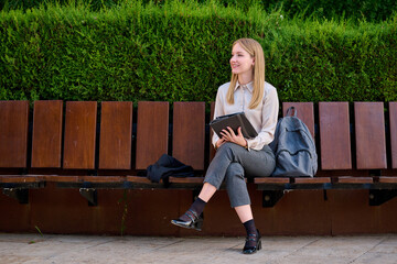 Young woman sitting on a bench in a park while holding a tablet