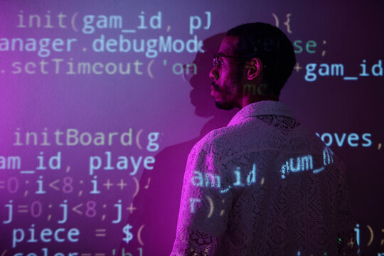 Side view of a person standing in a dimly lit room while observing computer code projected on a wall, providing a technological and introspective atmosphere