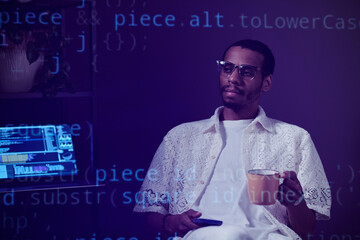 Man sitting in dimly lit workspace, holding cup of coffee while surrounded by digital coding...