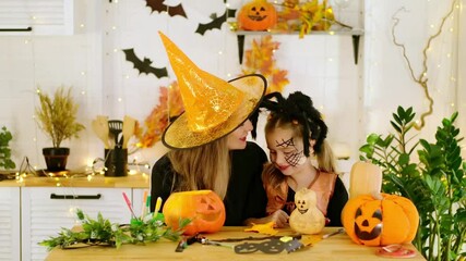 Halloween, a happy family a mother with a baby girl in festive costumes and makeup is preparing for Halloween at home in the kitchen by coloring and carving pumpkins and scaring everyone