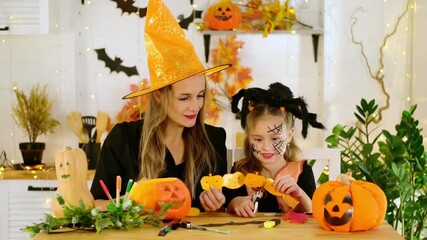 Halloween, a happy family a mother with a baby girl in festive costumes and makeup is preparing for Halloween at home in the kitchen by coloring and carving pumpkins and scaring everyone