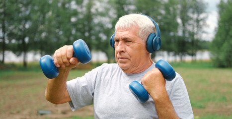 Fototapeta premium Old man exercising with dumbbells outside. Physical therapy. Elderly 60s male working out. Active sporty gray haired man standing with weights. Older person doing fitness outdoors. People, gym, sport