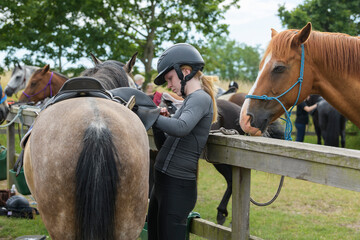 Young woman wearing equestrian helmet putting saddle on horse at paddock