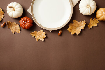 Autumn table setting with white plate and seasonal decor including pumpkins, acorns, and leaves on a brown backdrop. Perfect for Thanksgiving and fall dining visuals