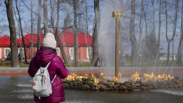Girl wearing a pink jacket and a white beanie is enjoying the beauty of the famous samson fountain in peterhof lower park, with its water jets gushing up high