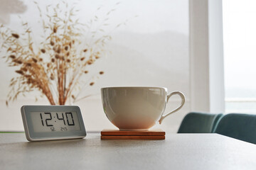 A digital clock with a white cup on a table. The clock shows 12:40 and the cup is on a wooden coaster.